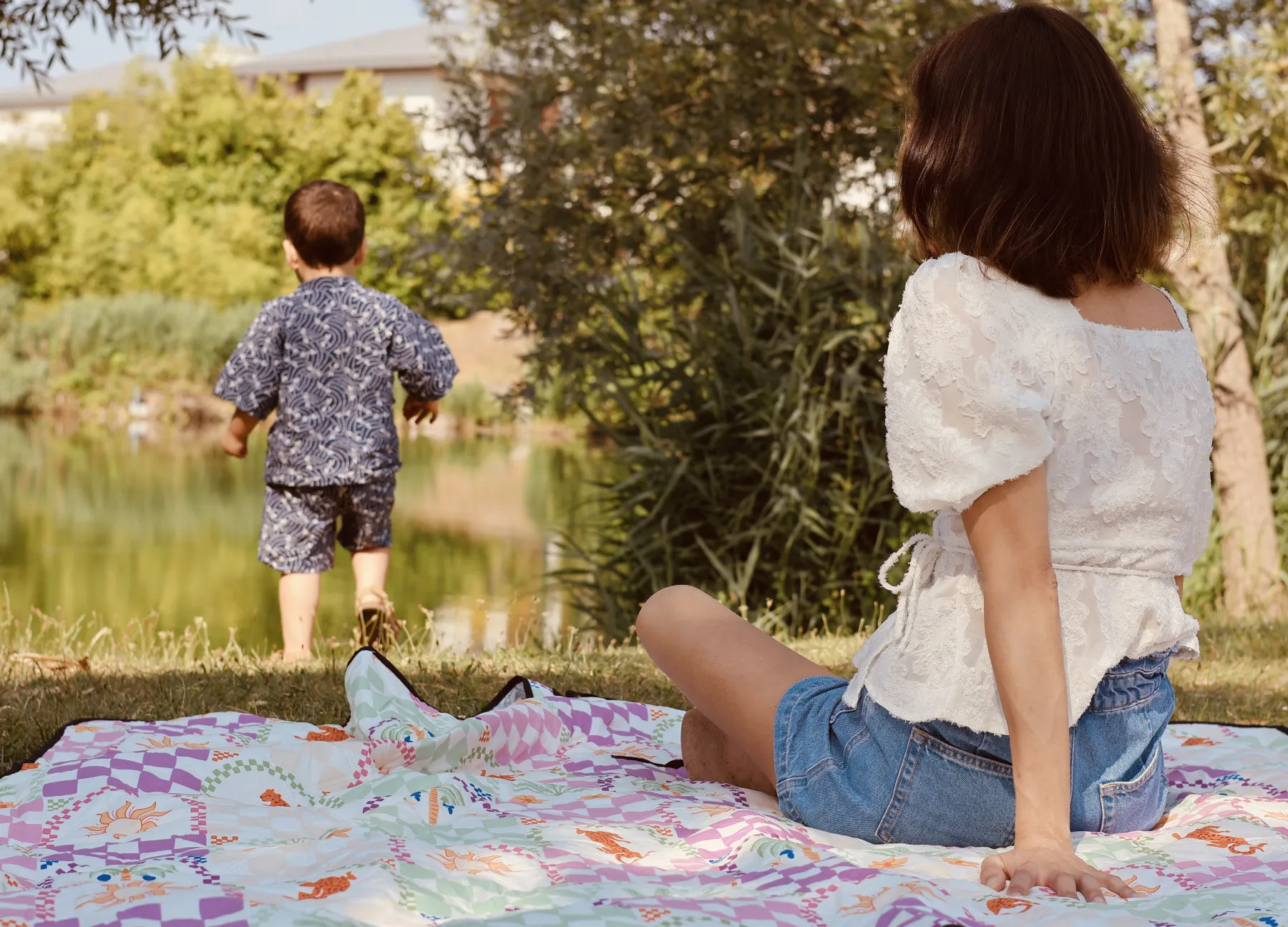 Une maman sur une couverture regarde son fils marcher vers l'étang
