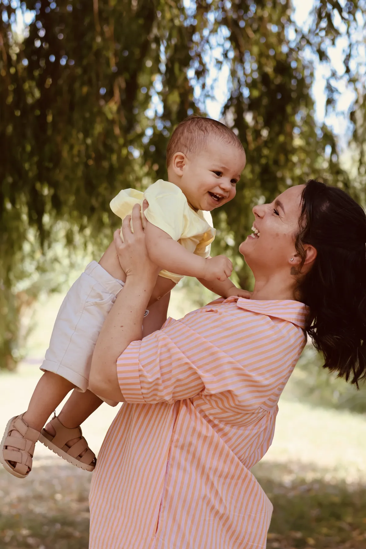 Une maman soulève son bébé rieur sous un saule pleureur