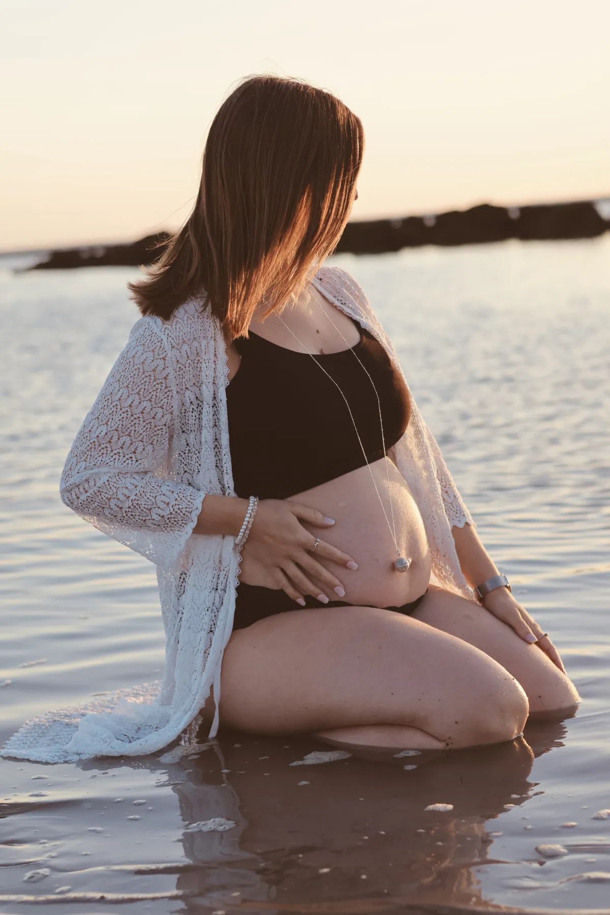 Future maman agenouillée dans l'eau au coucher du soleil, les mains sur son ventre