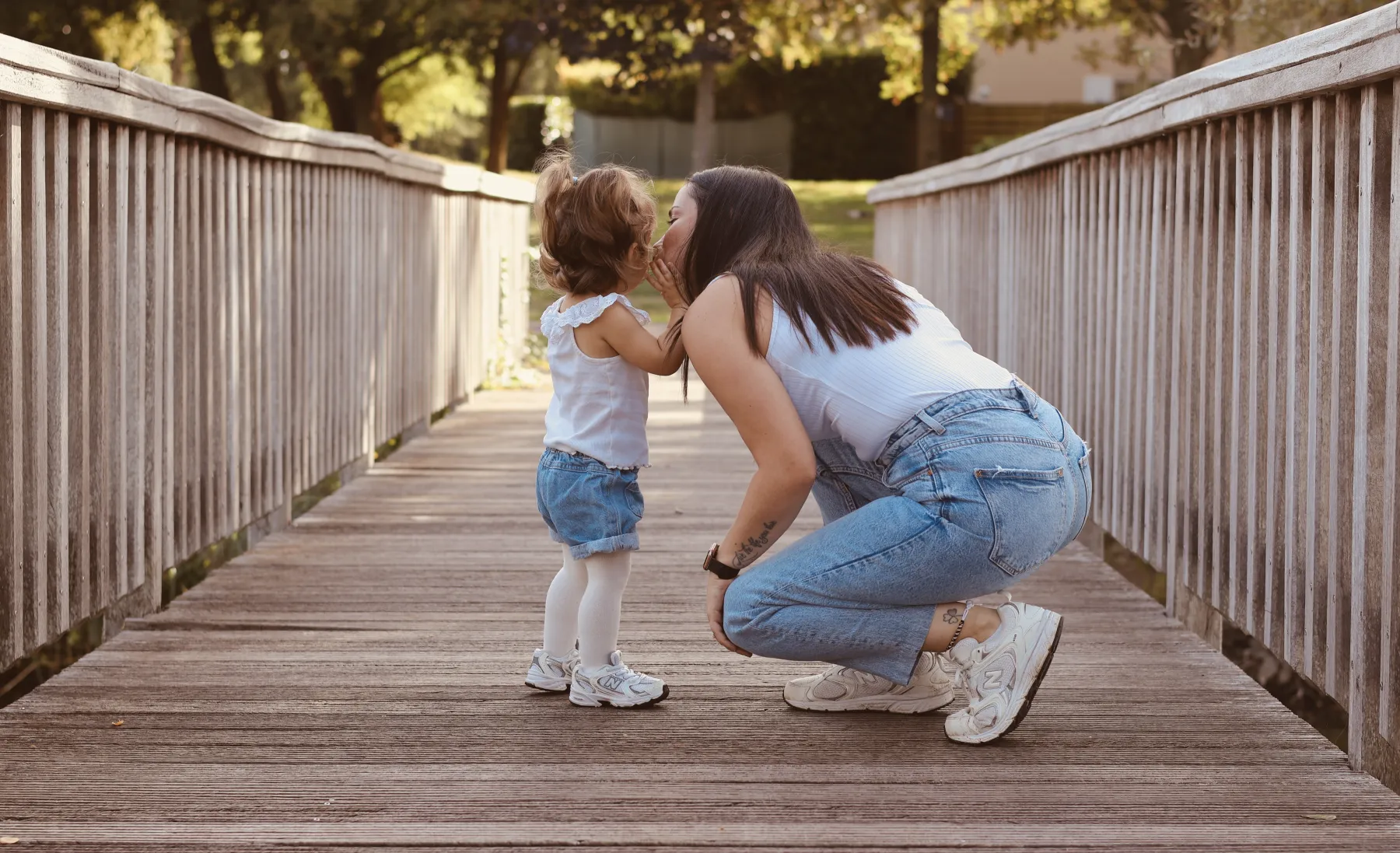 Une maman accroupie fait un bisou à sa petite fille sur un pont en bois