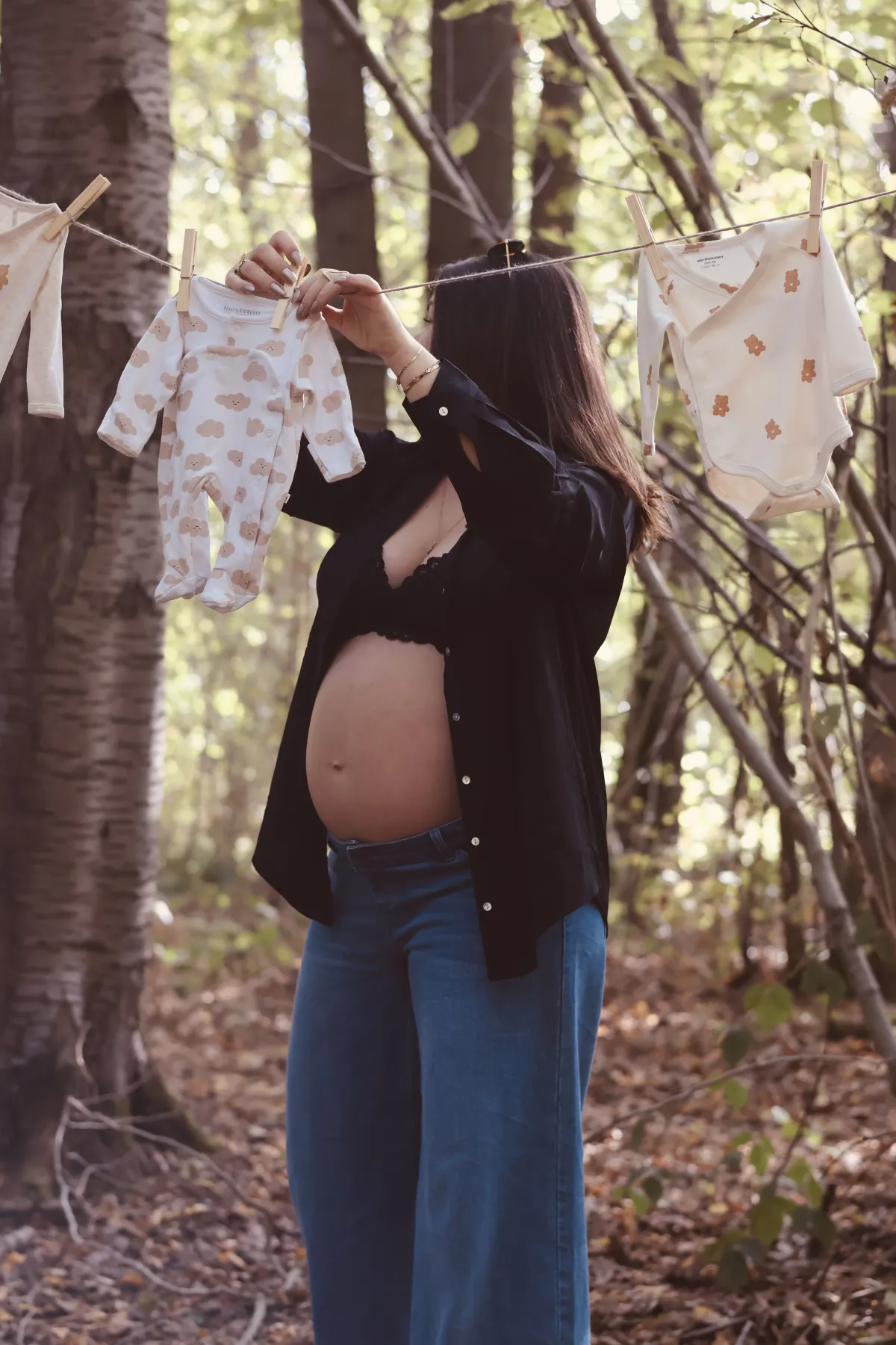 Future maman au ventre nu étend des petits bodies sur un fil, en forêt