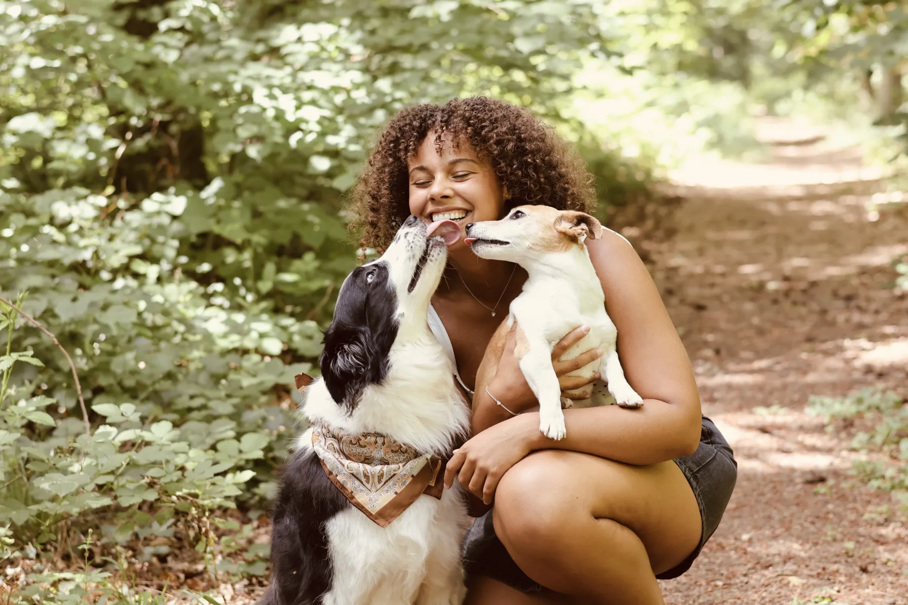 Jeune femme souriante embrassée par ses deux chiens sur un sentier